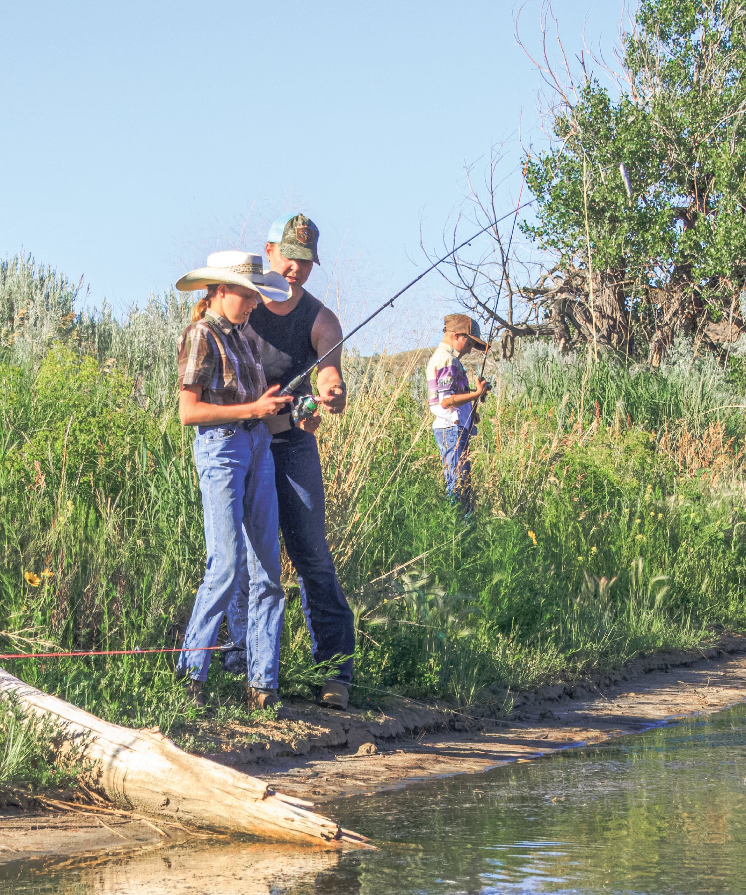 Three people fishing by a river with trees and clear sky in the background
