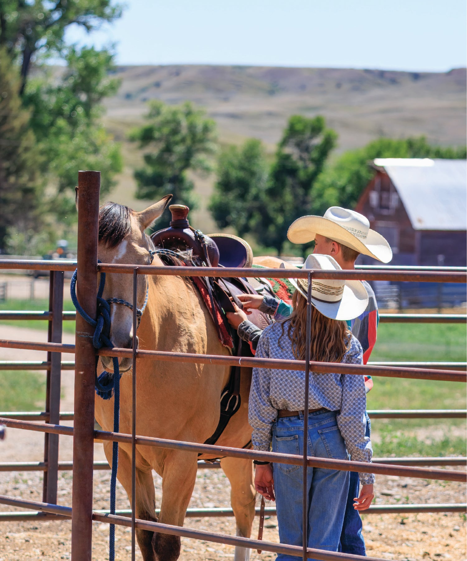 Two children in cowboy hats standing next to a horse in a fenced area with mountains in the background.