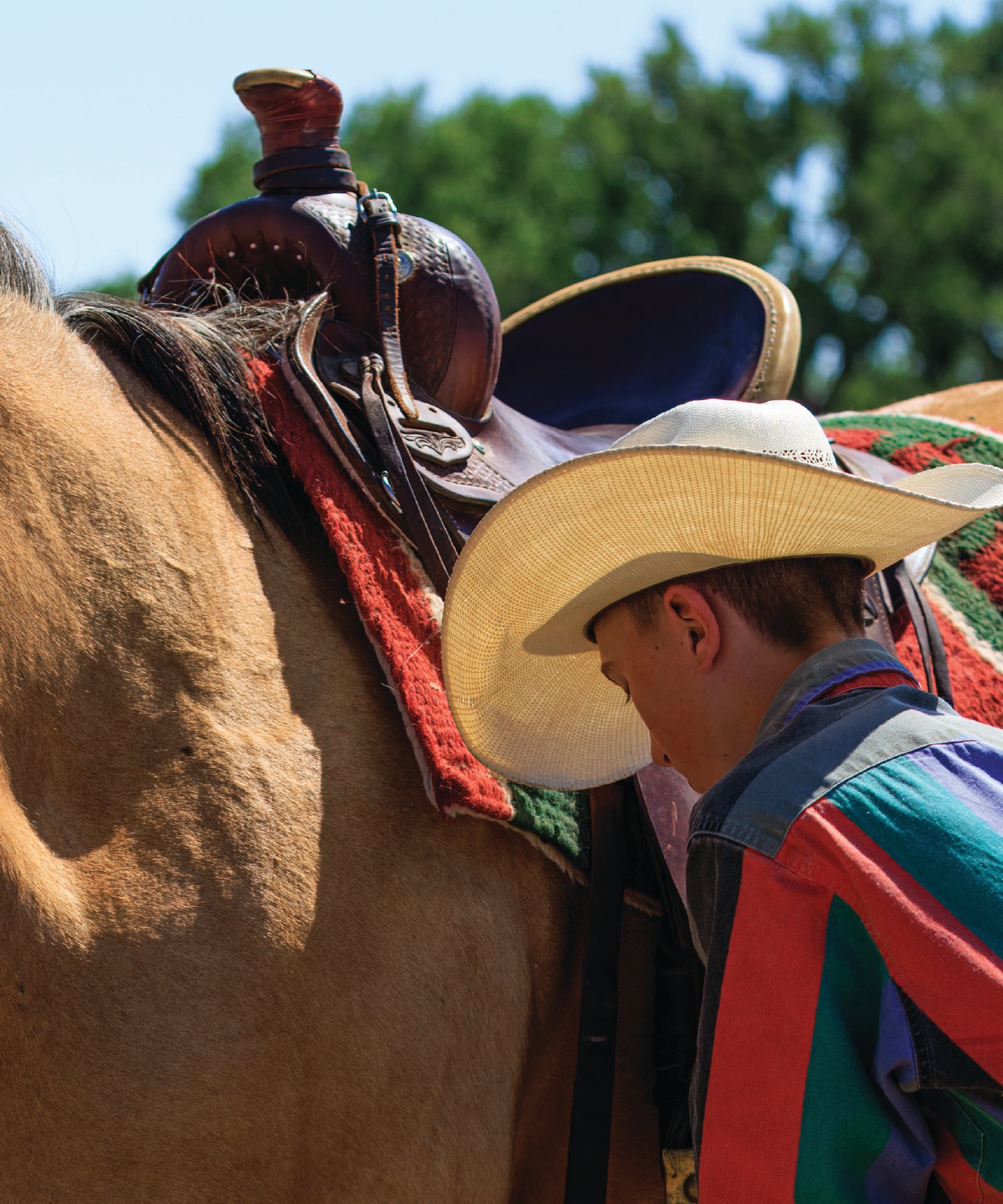 Person in a colorful outfit with a cowboy hat standing next to a horse with a saddle.