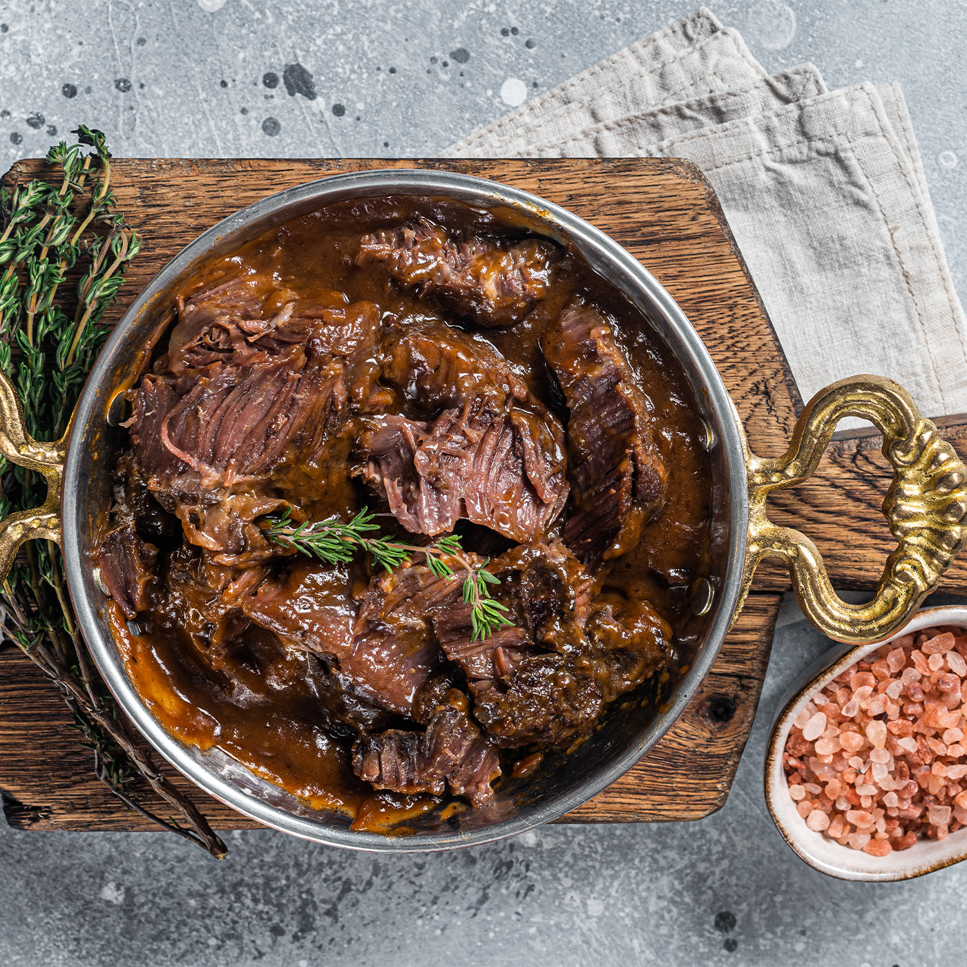 Braised beef in a metal pot on a wooden board with herbs and salt.
