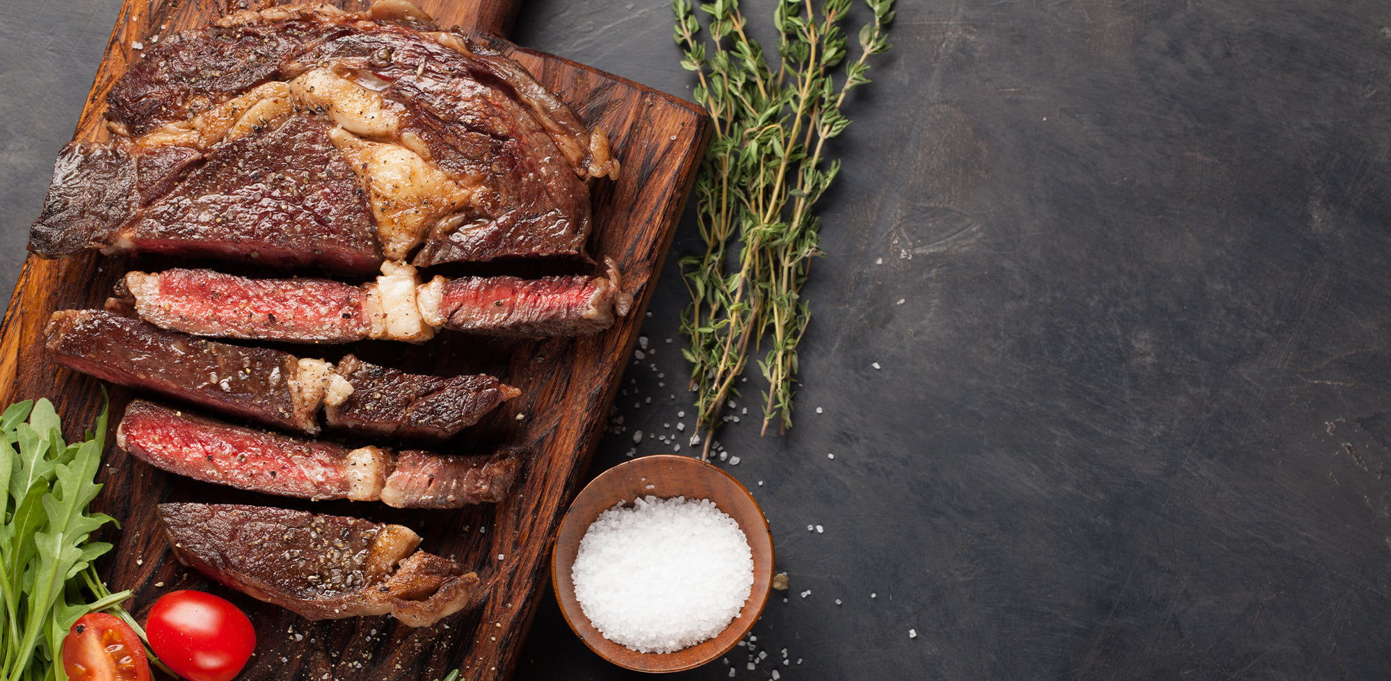 Sliced cured meat on a wooden board with herbs and salt on a dark surface