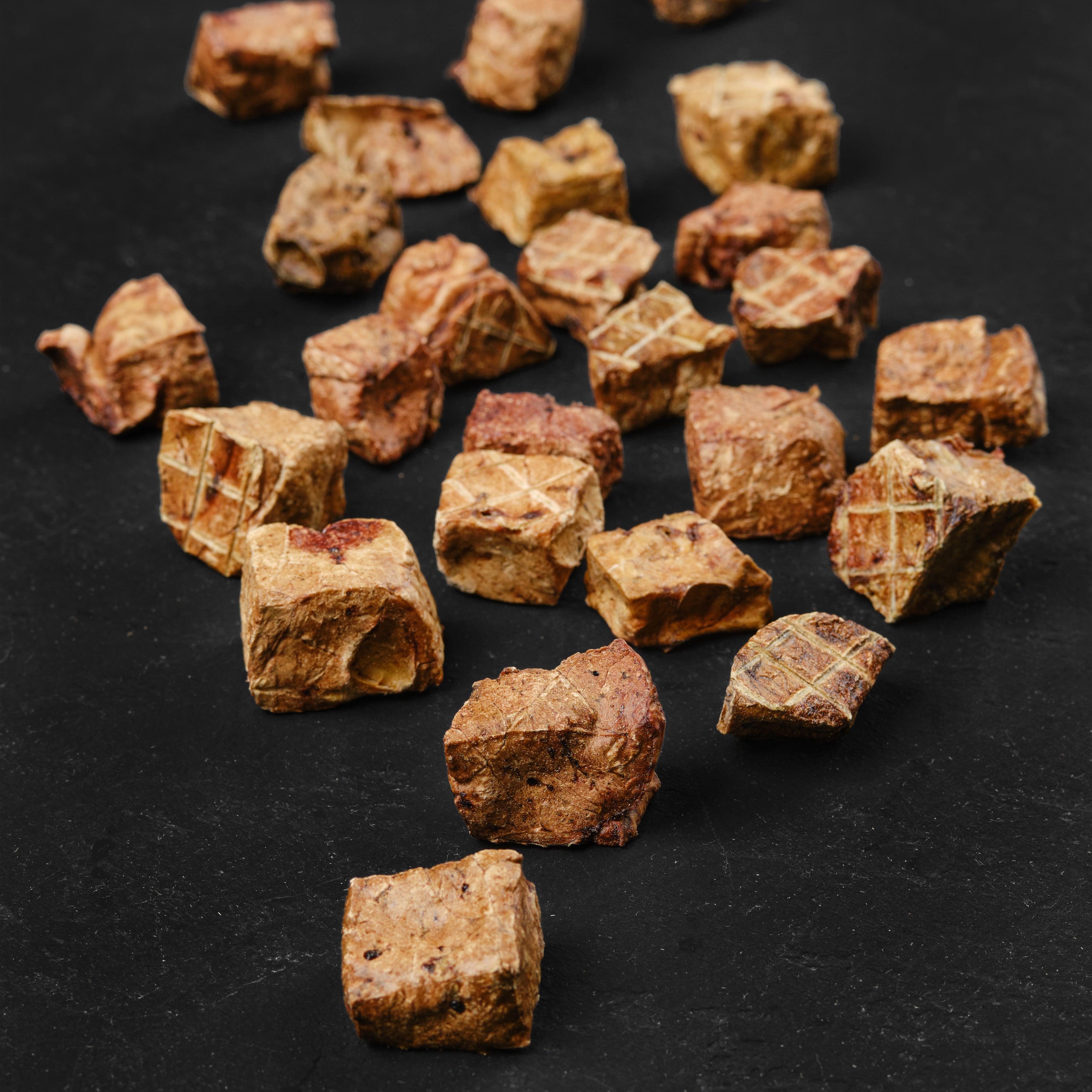 Dehydrated dog treats scattered on a black surface, resembling small, irregularly shaped cubes with a brown color.