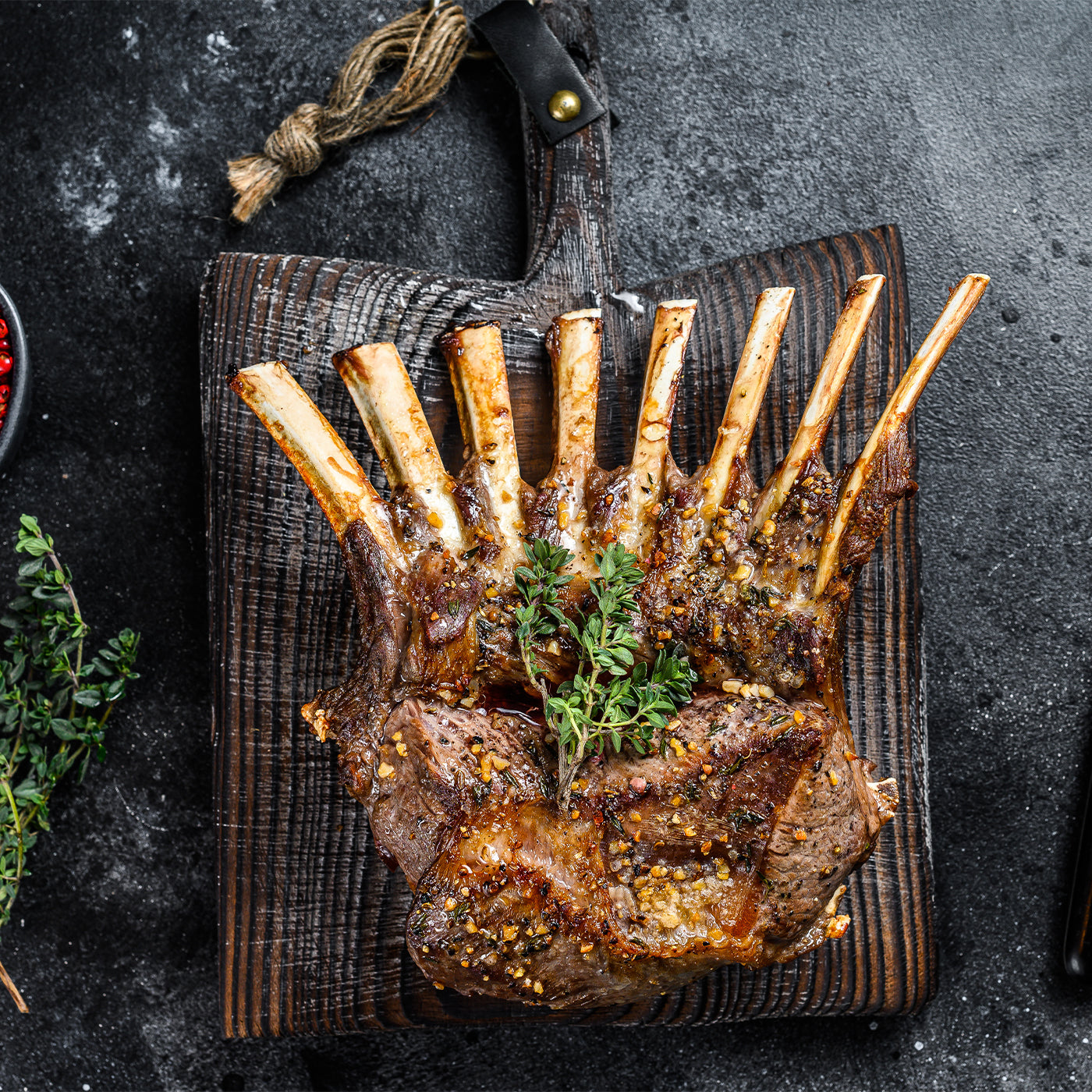 Grilled rack of lamb on a wooden cutting board with herbs on a dark surface